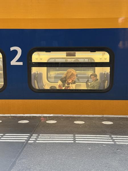 The photo captures a moment at Stationsplein, Centrum, Amsterdam, showing two passengers inside a bright yellow and blue train